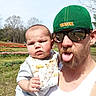 baby, beard, bib, cap, casual_clothing, child, father, flowers, grass, holding, man, outdoor, portrait, selfie_reflection, sky, sunglasses, sunlight, tongue_out, trees, tulip_field