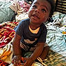 baby, toddler, child, indoor, bed, blanket, clothing, footwear, curly_hair, person, playing, striped_shorts, blue_shirt, room, messy, soft_toys, hands, looking_up, cute, casual