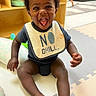 baby, child, bib, smiling, happy, indoor, playmat, toys, floor, sitting, clothing, person, infant, cute, expression, mouth_open, dark_skin, short_hair, room, furniture