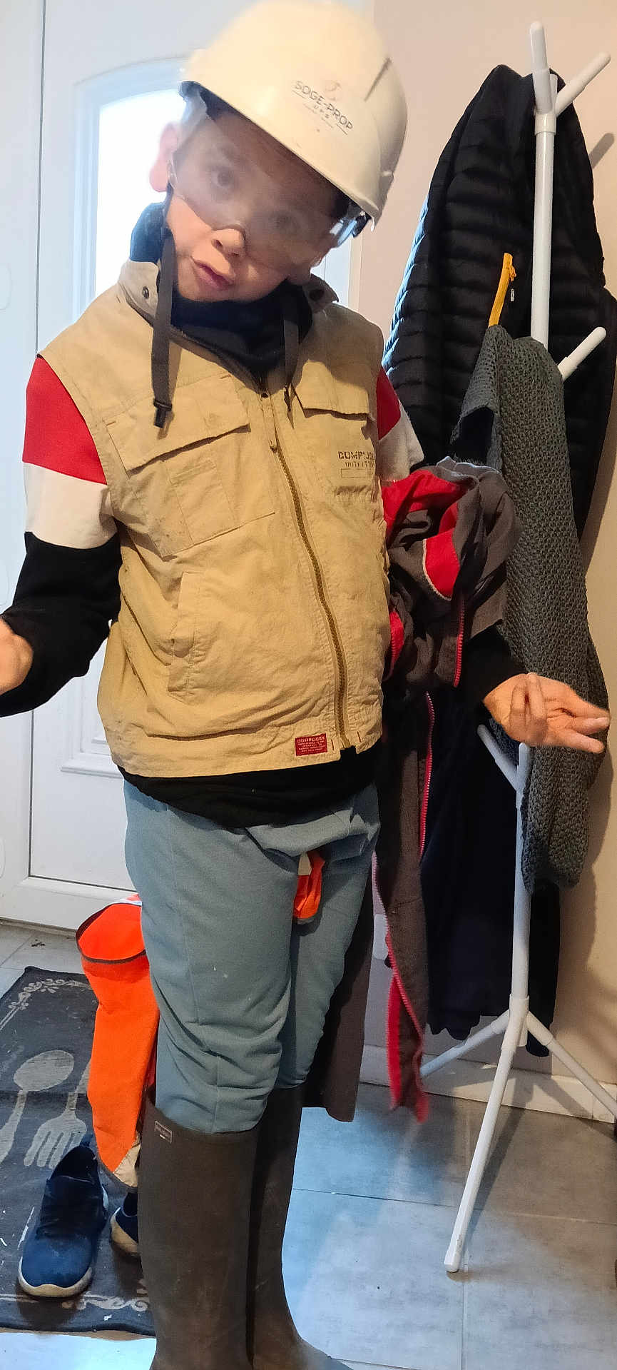 Mathis joined the competition — help win amazing prizes! child, kid, helmet, safety_goggles, vest, jacket, coat_rack, boots, rubber_boots, pants, shoes, mat, door, hallway, indoor, playful, silly_face, protective_gear, standing, entrance_mat