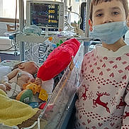 Mathis participe au concours pour gagner de l'argent avec cette photo : child, infant, baby, sibling, hospital_room, medical_monitor, incubator, hospital_crib, surgical_mask, sweater, reindeer_sweater, iv_bag, tubing, plush_toy, blanket, pillow, window, device_screen, visitor, medical_equipment