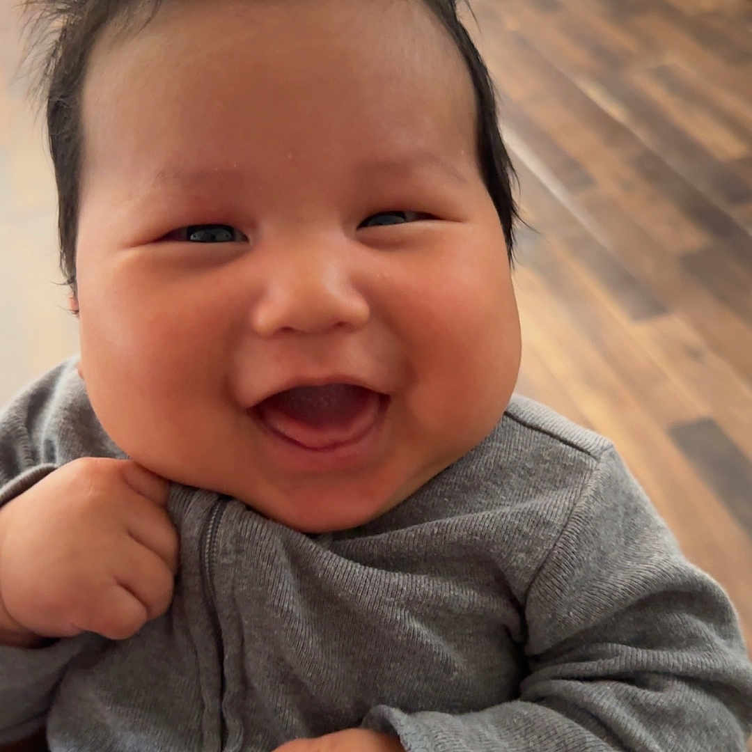 Jacoby is registered to the contest to win money with this photo: baby, smile, happy, infant, child, face, clothing, gray_outfit, indoor, wooden_floor, hand, cheeks, cute, person, sitting, cozy, young_child, portrait, expression, toy