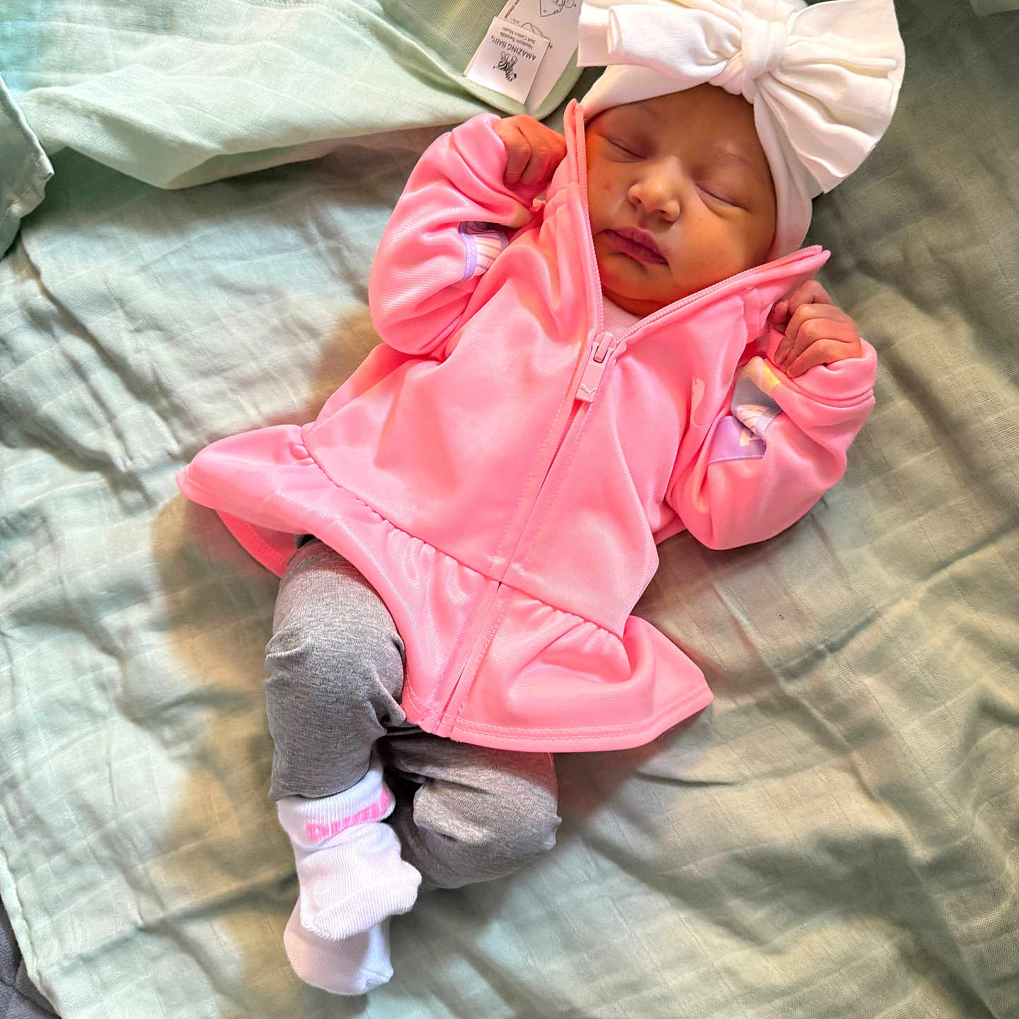 Sarayah is registered to the contest to win money with this photo: baby, blanket, child, clothing, cozy, cute, gray_pants, headwear, indoors, infant, nap, newborn, peaceful, pink_jacket, resting, sleeping, small_hands, socks, soft_fabric, white_bow