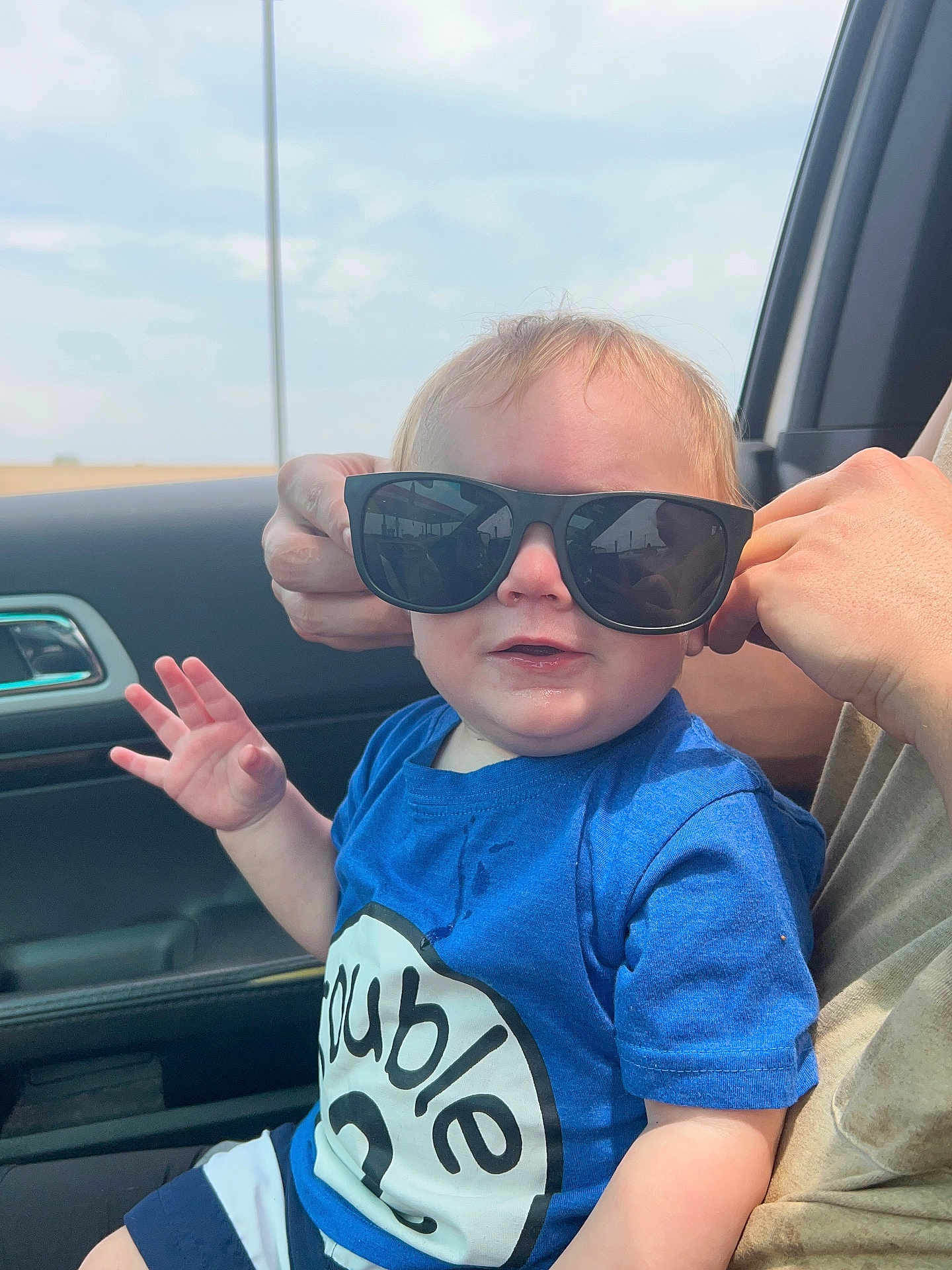 Gunnar is registered to the contest to win money with this photo: toddler, child, sunglasses, blue_shirt, car_interior, hand, adult_hand, smile, face, window, sky, seat, clothing, shorts, light_skin, person, casual, sitting, playful, sunlight