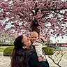 affection, baby, bicycle_rack, blossom_tree, bush, cherry_blossom, coat, face, flowers, holding, infant, jacket, kiss, mother, outdoor, park, pavement, pink, portrait, spring