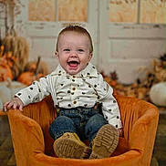 Cannon is registered to the contest to win money with this photo: baby, child, smile, happy, suspenders, jeans, orange_chair, indoor, autumn, pumpkins, background, plush_chair, cute, portrait, sitting, person, floor, shirt, footwear, decor