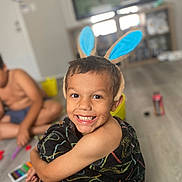 Niko is registered to the contest to win money with this photo: child, boy, smile, bunny_ears, blanket, indoor, floor, toy, play, living_room, television, sitting, happy, person, furniture, blurred_background, phone, casual, home, cute