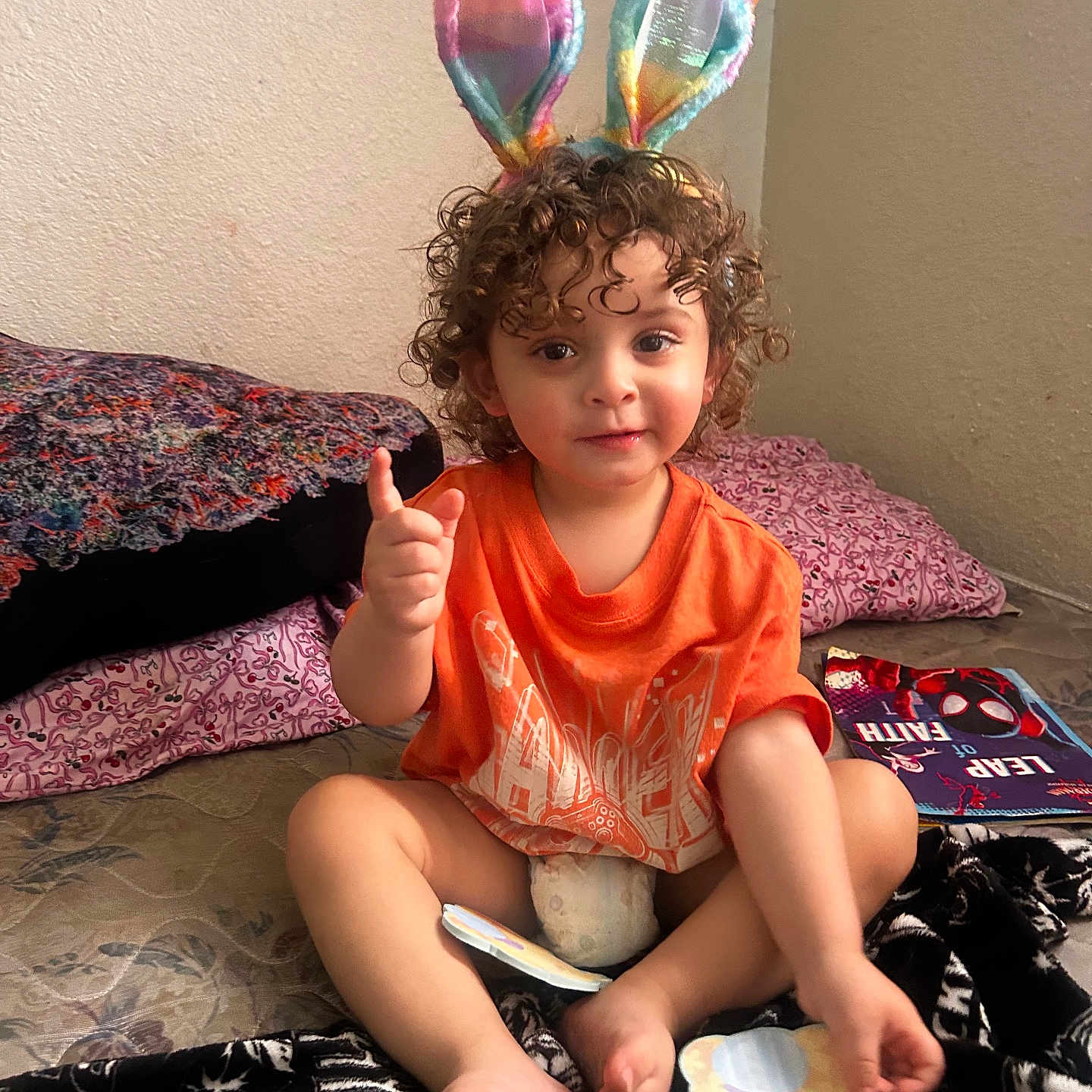 King is registered to the contest to win money with this photo: child, toddler, bunny_ears, orange_shirt, curly_hair, bed, pillow, blanket, paper_cutout, indoor, smiling, cute, playful, person, feet, hand, wall, colorful, expression, seated