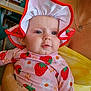 baby, child, person, infant, hat, strawberry_pattern, flower_pattern, clothing, drool, hand, fan, yellow_top, indoor, table, chair, window, reflection, face, portrait, holding