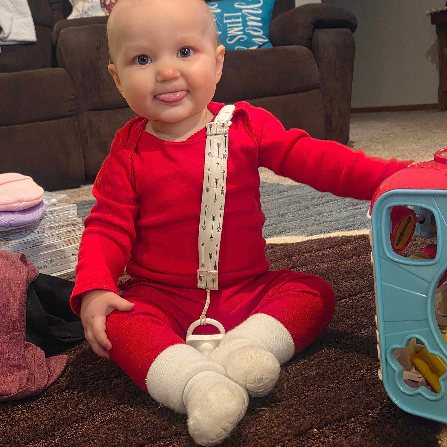 Macie Rae joined the competition — help win amazing prizes! baby, carpet, carpeted_floor, child, couch, cute, floor, furniture, indoors, living_room, pacifier, person, pillow, playful, red_clothing, sitting, smiling, tongue_out, toy, white_socks