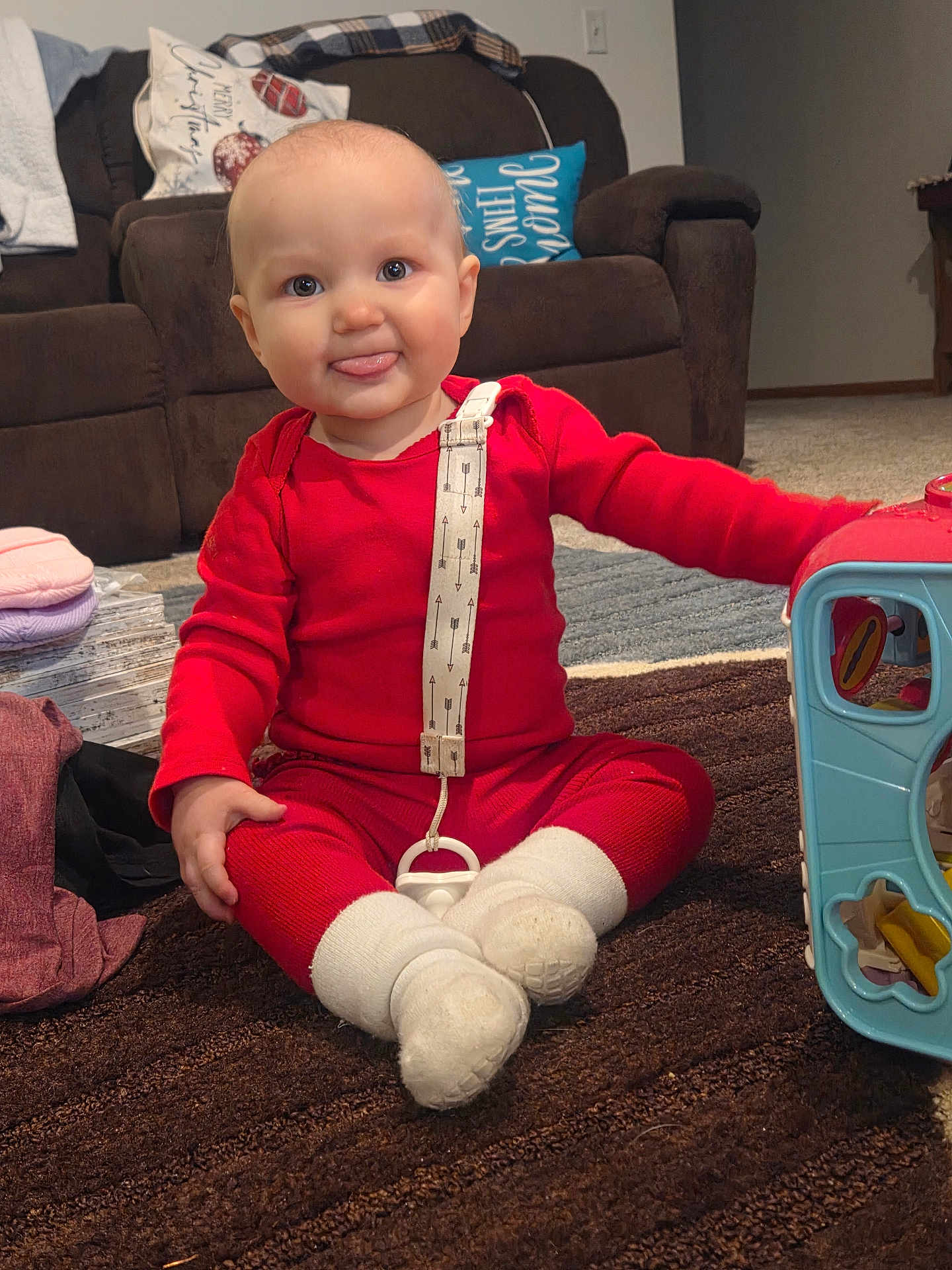 Macie Rae joined the competition — help win amazing prizes! baby, child, red_clothing, tongue_out, sitting, indoors, toy, carpet, living_room, couch, pillow, pacifier, white_socks, playful, cute, smiling, floor, carpeted_floor, furniture, person