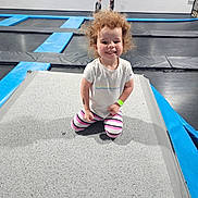 Paityn joined the competition — help win amazing prizes! child, toddler, curly_hair, smiling, white_tshirt, striped_leggings, trampoline, playground, indoor, recreation, fun, happy, kneeling, blurred_background, kids, play, sport, exercise, wristband, gym