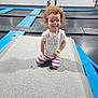 child, toddler, curly_hair, smiling, white_tshirt, striped_leggings, trampoline, playground, indoor, recreation, fun, happy, kneeling, blurred_background, kids, play, sport, exercise, wristband, gym