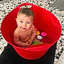 baby, bath_toys, black_rug, child, cute, floor, happy, head, indoor, person, playful, red_bucket, sitting, skin, smiling, tile_floor, toy_duck, toy_fish, toy_pig, water