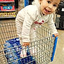 Mia is registered to the contest to win money with this photo: child, toddler, shopping_cart, supermarket, store_interior, smiling, playful, water_bottles, packaged_goods, shoes, hoodie, tracksuit, aisle, people, shelves, snacks, floor, shopping_trip, portrait, candid