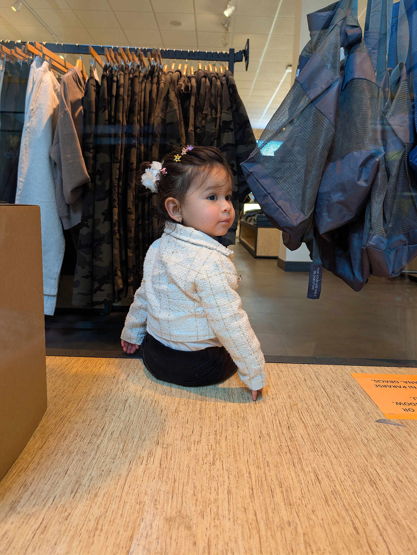 Mia is registered to the contest to win money with this photo: toddler, child, clothing_store, clothes_rack, hangers, jackets, camouflage_pattern, shopping_bags, retail, sitting, looking_back, hair_accessories, earrings, jacket, wooden_floor, display, glass_reflection, box, floor_sign, portrait