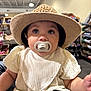 baby, hat, pacifier, indoor, child, curious, white_clothing, sitting, wide_eyes, infant, person, store, shelves, boxes, cute, portrait, face, headwear, young_child, casual