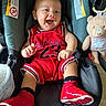 baby, child, infant, smiling, laughing, car_seat, teddy_bear, red_outfit, basketball_jersey, sneakers, shoes, portrait, indoor, seated, happy, toy, vehicle, face, close_up, playful