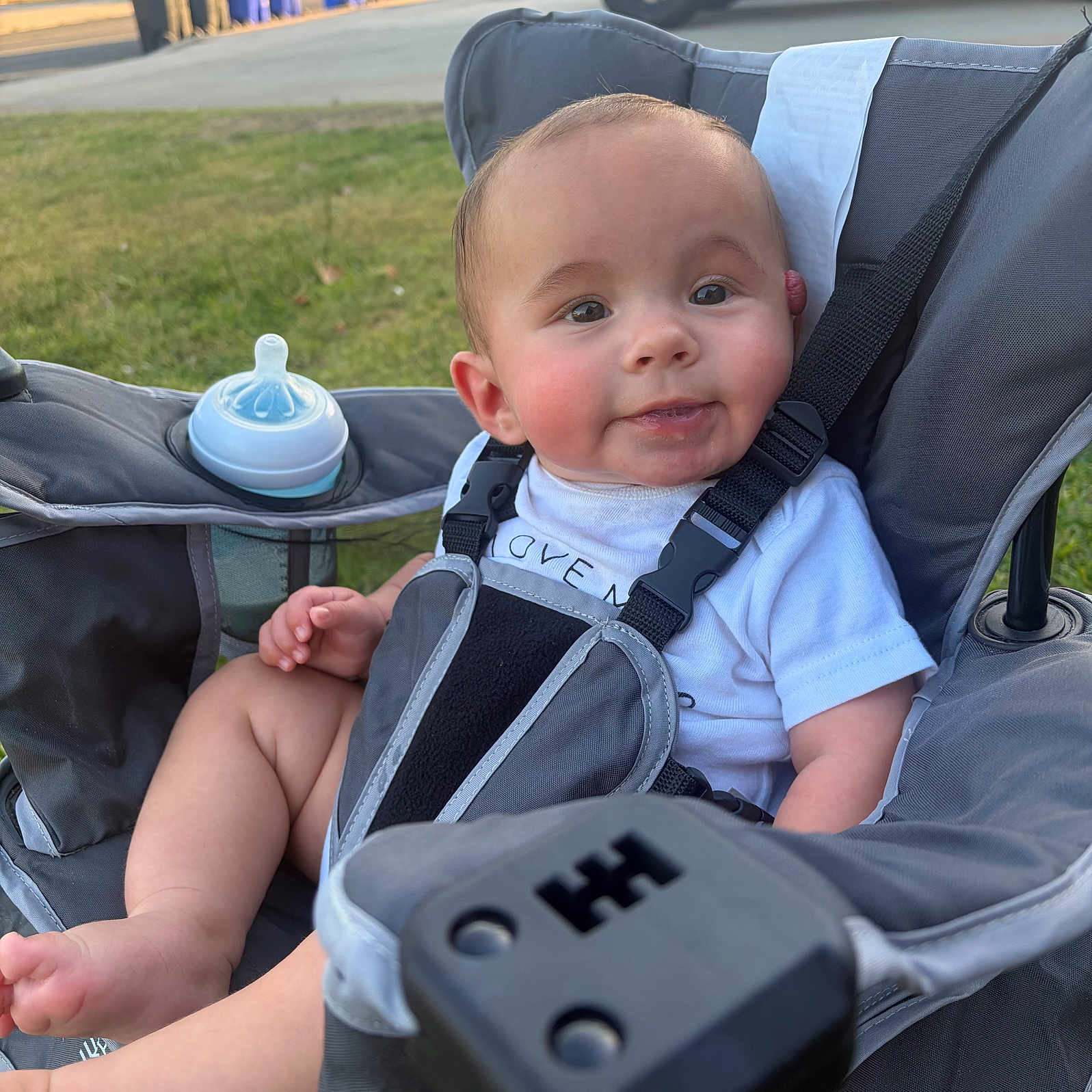 Sonny Cazares is registered to the contest to win money with this photo: baby, stroller, bottle, outdoor, child, infant, cute, seatbelt, grass, sidewalk, car, trash_bins, sunlight, white_shirt, face, smile, person, legs, hand, parked_vehicle