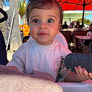 Eyden a rejoint le concours — aidez-le/la à gagner de superbes lots ! toddler, child, high_chair, toy, phone, outdoor, cafe, umbrella, red_umbrella, palm_tree, sunny, daylight, clothing, person, face, expression, hand, table, seat, background