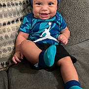 Jakobee is registered to the contest to win money with this photo: baby, child, smile, blue_clothing, hat, socks, couch, cushion, indoor, portrait, happy, person, infant, sporty_outfit, seated, cute, small_child, face, one_person, cozy