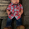 baby, child, plaid_shirt, jeans, brown_shoes, couch, cushions, patterned_cushion, indoor, seated, curious_expression, wide_eyes, casual_clothing, portrait, infant, furniture, young_child, comfortable, home, person