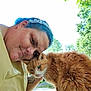 person, cat, orange_cat, outdoor, greenery, trees, daylight, affection, selfie, headband, tattoo, fur, pet, nature, closeup, friendship, smile, collar, bench, cuddling