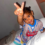 Blossom is registered to the contest to win money with this photo: child, smiling, peace_sign, colorful_clothing, striped_pants, bed, blanket, indoor, portrait, happy, person, young_child, cute, playful, hand_gesture, casual_wear, hair_buns, bedroom, cozy, relaxed