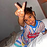 child, smiling, peace_sign, colorful_clothing, striped_pants, bed, blanket, indoor, portrait, happy, person, young_child, cute, playful, hand_gesture, casual_wear, hair_buns, bedroom, cozy, relaxed