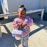 child, girl, smiling, outdoor, sunlight, toy, handbag, pink_sweater, colorful_clothing, sandals, happy, cute, playful, standing, shadow, concrete, background_wall, hair_bun, casual, daylight