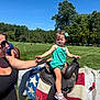 Avery is registered to the contest to win money with this photo: child, toddler, pony, horse, saddle, saddle_blanket, outdoor, grass, tree, woman, sunglasses, blue_sky, sunny, greenery, holding, smiling, clothing, footwear, hair_bow, recreation