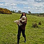 person, dog, grass, field, clouds, sky, outdoor, nature, rural, happy, holding, green, landscape, fur, boots, casual_clothing, trees, shrubbery, leisure, daytime