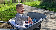 Aaron a rejoint le concours — aidez-le/la à gagner de superbes lots ! toddler, child, wheelbarrow, blanket, road, grass, trees, sky, clouds, wind_turbine, greenery, sunlight, outdoor, nature, happy, smile, casual_clothing, daytime, rural, transport
