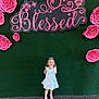 Gracelyn is registered to the contest to win money with this photo: child, girl, white_dress, pink_shoes, smiling, standing, green_wall, pink_flowers, decor, sign, text, happy, portrait, floor, cute, background, fashion, person, indoor, casual