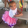 toddler, child, smiling, colorful_beads, pink_tutu, white_shirt, outdoor, picnic_table, concrete_floor, bench, person, sneakers, shorts, casual_clothing, festive, ribbons, colorful_fabric, happy, daylight, playful