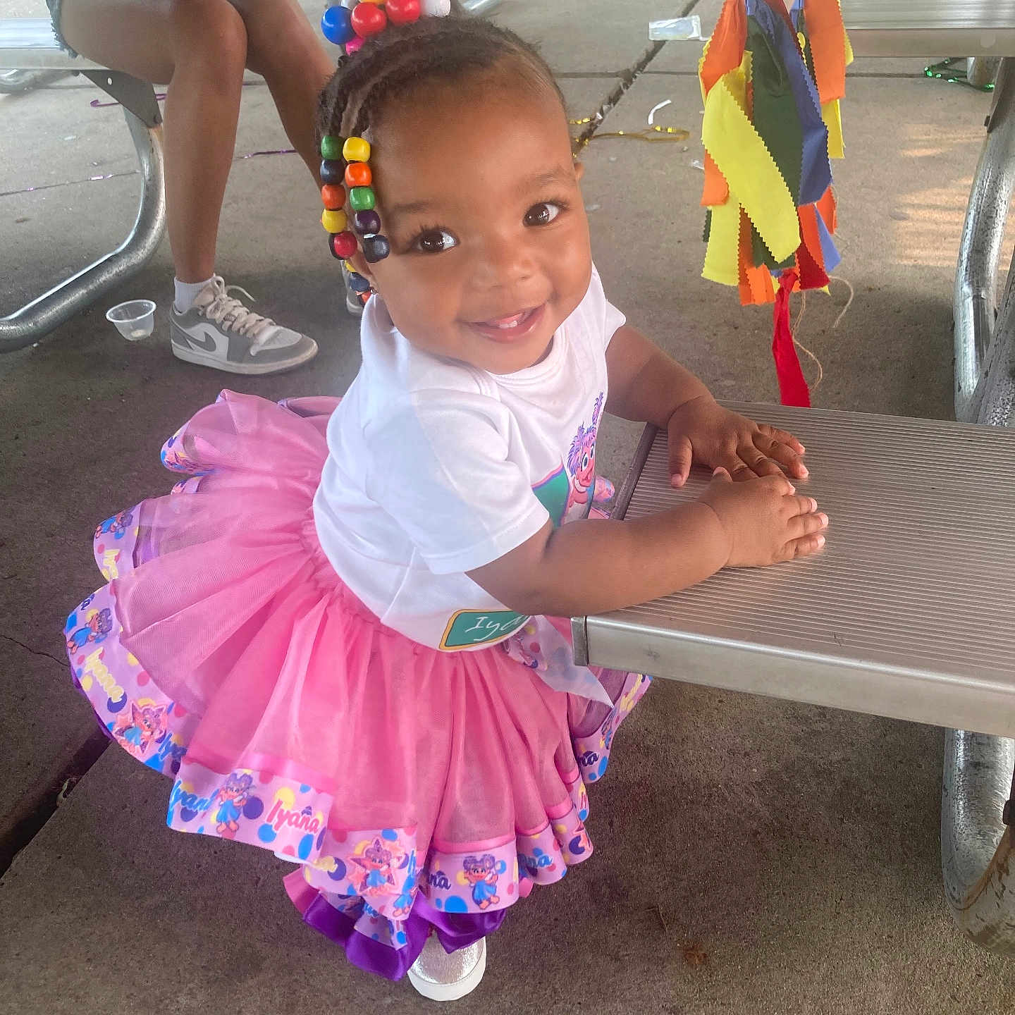 Yaya joined the competition — help win amazing prizes! toddler, child, smiling, colorful_beads, pink_tutu, white_shirt, outdoor, picnic_table, concrete_floor, bench, person, sneakers, shorts, casual_clothing, festive, ribbons, colorful_fabric, happy, daylight, playful