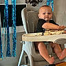 baby, child, high_chair, birthday, cake, frosting, messy, party, streamers, blue_decorations, balloons, indoor, sitting, face, portrait, black_shirt, barefoot, celebration, home, happy
