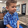 child, boy, curly_hair, blue_shirt, striped_shirt, blue_shorts, sitting, indoor, barbershop, salon, chair, mirror, ceiling_lights, waiting, portrait, casual_clothing, young, cute, person, haircut