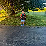 child, boy, smiling, preschool, first_day, sign, backpack, tree, grass, driveway, sunlight, outdoor, nature, happy, casual_clothing, shorts, sneakers, greenery, morning, portrait