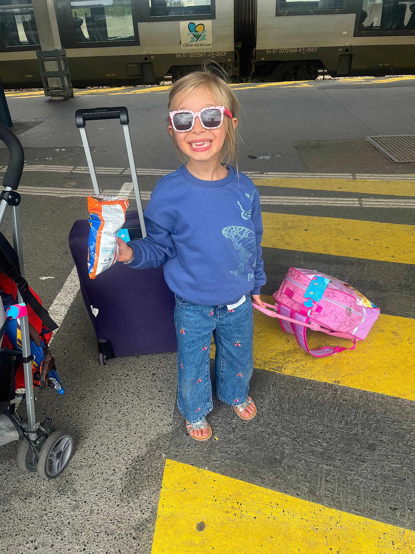Eloïse a rejoint le concours — aidez-le/la à gagner de superbes lots ! child, girl, sunglasses, blue_sweatshirt, butterfly, jeans, sandals, luggage, pink_suitcase, train_station, train, platform, snack, smile, travel, waiting, outdoor, daylight, road_markings, stroller