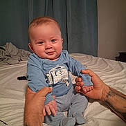 Aaron joined the competition — help win amazing prizes! baby, child, infant, blue_clothing, tattoo, hands, bed, blanket, curtains, smile, sitting, person, indoor, cozy, remote_control, pajamas, cute, portrait, skin, face