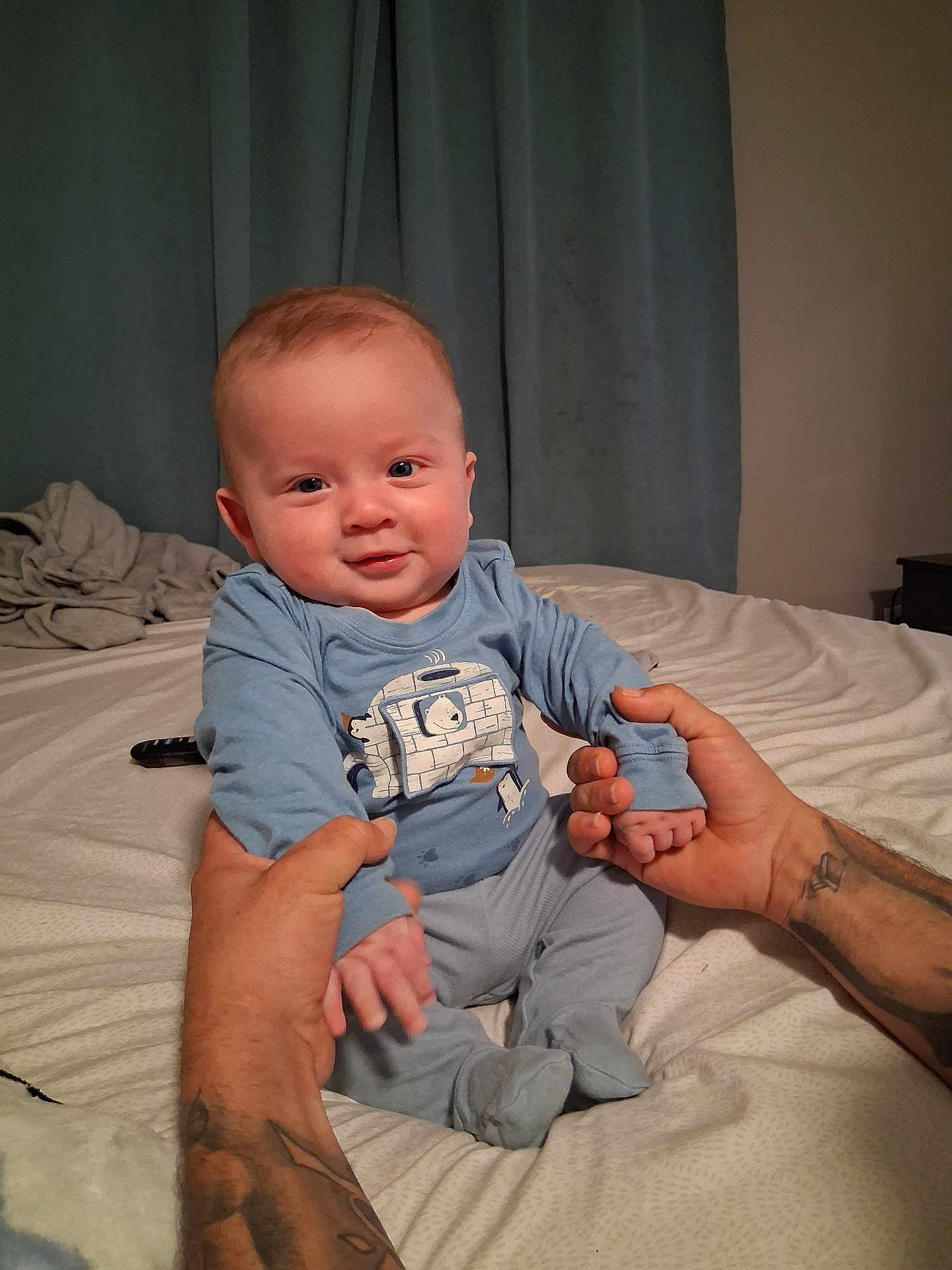 Aaron joined the competition — help win amazing prizes! baby, child, infant, blue_clothing, tattoo, hands, bed, blanket, curtains, smile, sitting, person, indoor, cozy, remote_control, pajamas, cute, portrait, skin, face