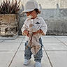 toddler, child, pacifier, plush_toy, cap, striped_pants, white_shoes, outdoor, plant, potted_plant, wall, tile_floor, cute, standing, young_child, clothing, casual, innocence, daylight, portrait