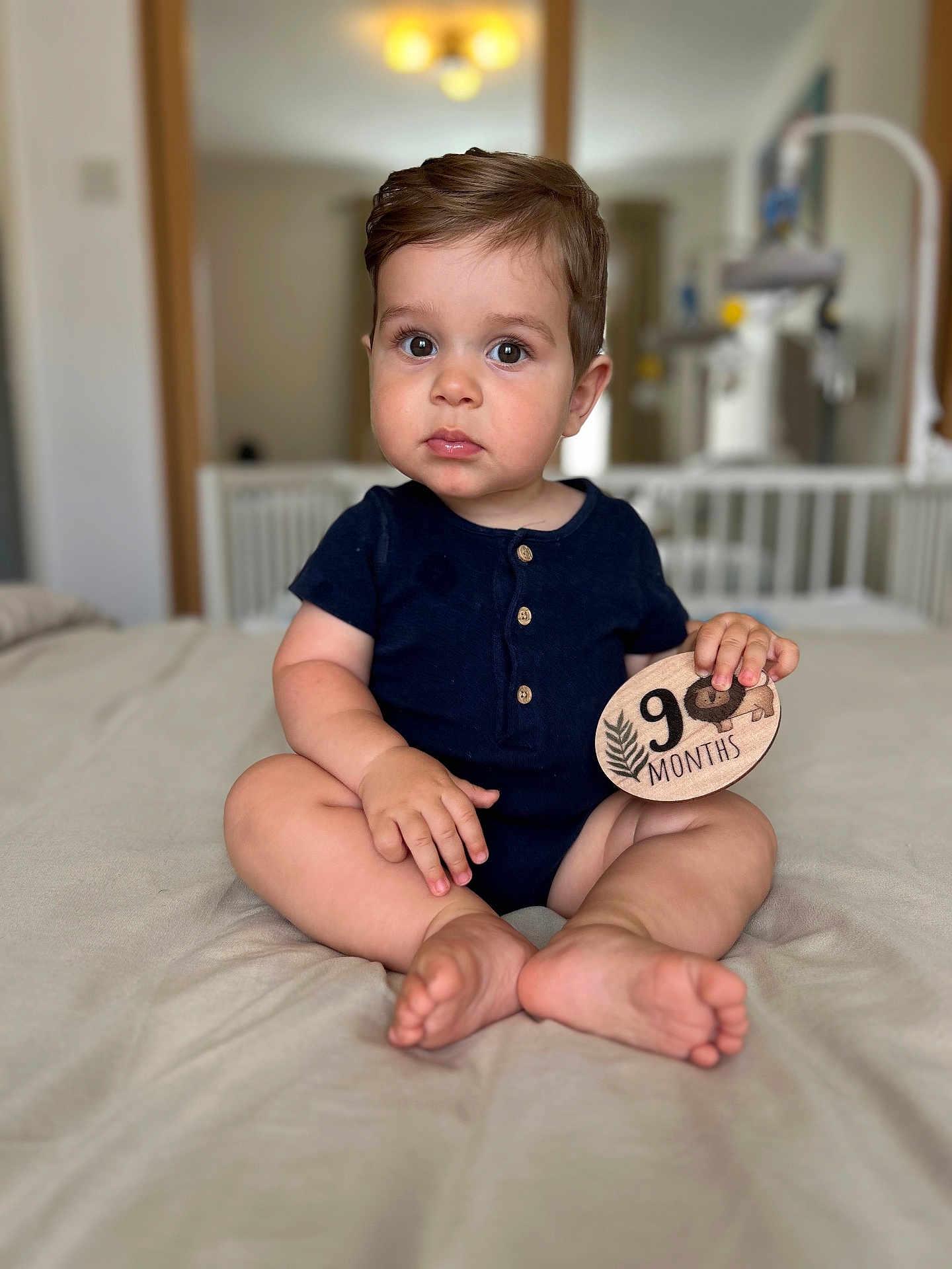 David is registered to the contest to win money with this photo: baby, infant, child, onesie, bed, plaque, 9_months, sitting, indoors, crib, wood, milestone, portrait, cute, chubby_cheeks, person, furniture, soft_lighting, hands, feet