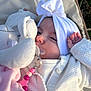 baby, infant, pacifier, headband, bow, blanket, sweater, plush_toy, pacifier_clip, hand, eyes, face, portrait, close_up, outdoors, stroller, grass, soft_texture, pink, white