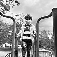 Ayden participe au concours pour gagner de l'argent avec cette photo : child, boy, playground, slide, jeans, jacket, sneakers, outdoor, black_and_white, portrait, handrails, trees, cloudy_sky, casual_clothing, young, person, fashion, urban, daytime, expression