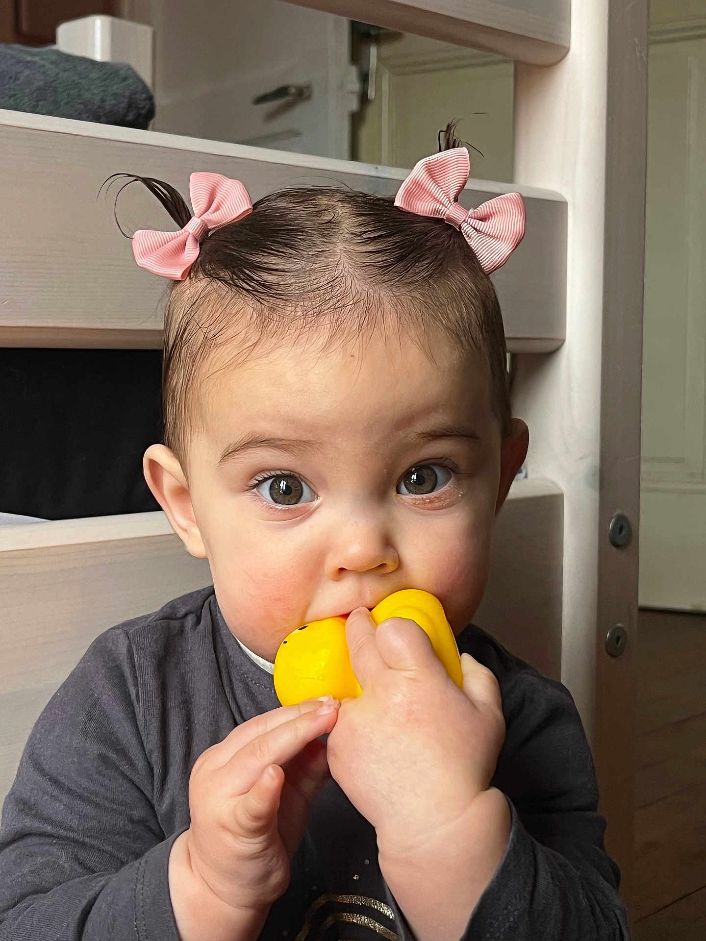 Esmée participe au concours pour gagner de l'argent avec cette photo : toddler, child, baby, toy, yellow_toy, pink_bows, hair_bows, indoor, wooden_furniture, curious, chewing, face, hands, cute, portrait, person, young_child, clothing, long_sleeves, expression