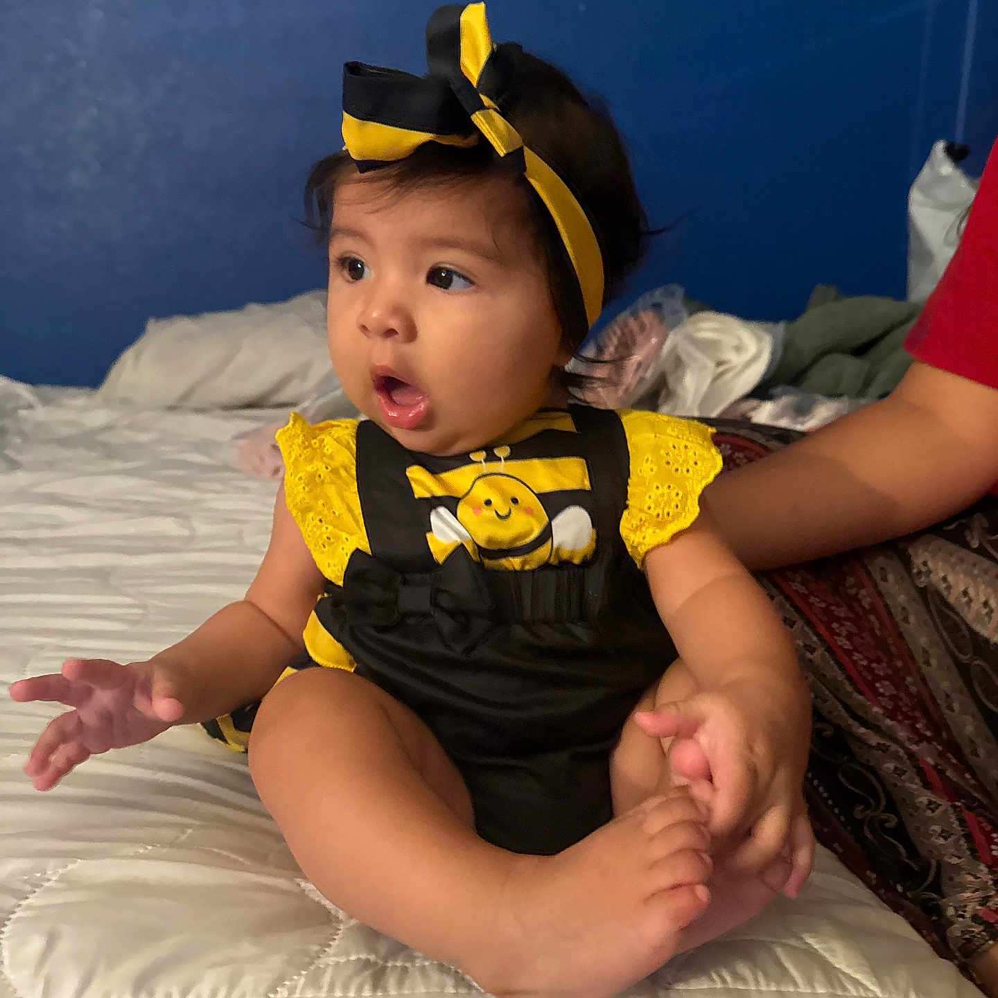 Camila is registered to the contest to win money with this photo: baby, bed, black, blue_wall, child, clothing, curious, cute, expression, foot, hand, headband, holding, indoor, infant, person, room, sitting, soft_light, yellow