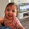 Aylanna is registered to the contest to win money with this photo: baby, child, closeup, curly_hair, cute, expression, face, floor, happy, holding, human, indoor, infant, living_room, modern, person, pink_clothing, portrait, smiling, television