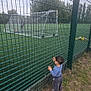 toddler, child, soccer_field, fence, grass, outdoor, sport, goalpost, playing_field, person, greenery, trees, overcast_sky, curious, young_child, casual_clothing, daytime, nature, recreation, activity